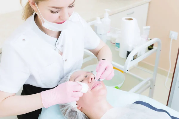 Un primo piano della procedura di pulizia nell'ufficio di cosmetologia. Le mani del cosmetologo in guanti rosa vengono rimosse dal viso di una giovane ragazza con una spugna che pulisce la maschera. — Foto Stock