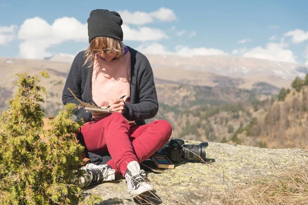 선글라스와 푸른 하늘에 대하여 산에 바위 야외에 앉아 모자를 입고 hipster 여자의 초상화. 프리랜서 디자이너 여행에 노트북에는 연필을 그립니다. — 스톡 사진