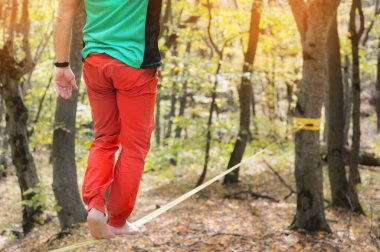 Arkadan bakan erkek ip cambazı sonbahar ormanında çıplak ayakla ip üzerinde dengede duruyor. Açık hava sporları konsepti ve yaşlanan insanların aktif hayatı