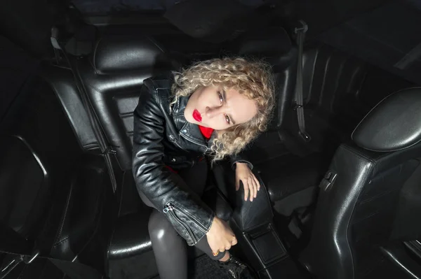 Estilo de moda de pantalla ancha joven y elegante chica atractiva con labios de color rojo brillante en una chaqueta de cuero y gafas de sol se sienta en el interior de cuero de un coche retro de los años 90 — Foto de Stock