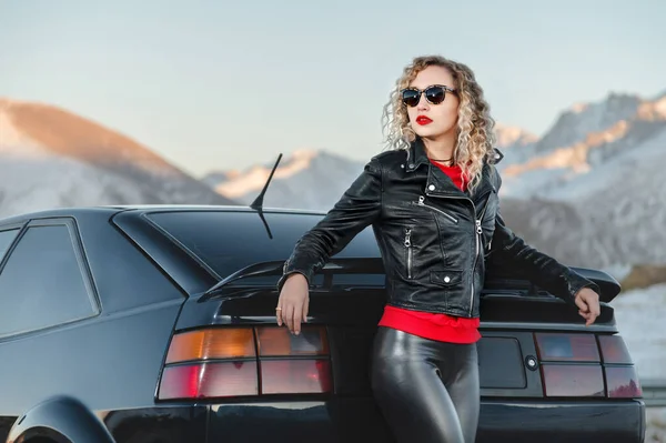 Moda styl szeroki kąt młoda stylowa atrakcyjna dziewczyna z jasnymi czerwonymi ustami w skórzanej kurtce i okularach stoi obok samochodu lat 90-tych retro. o przyrodzie w górach o zachodzie słońca — Zdjęcie stockowe