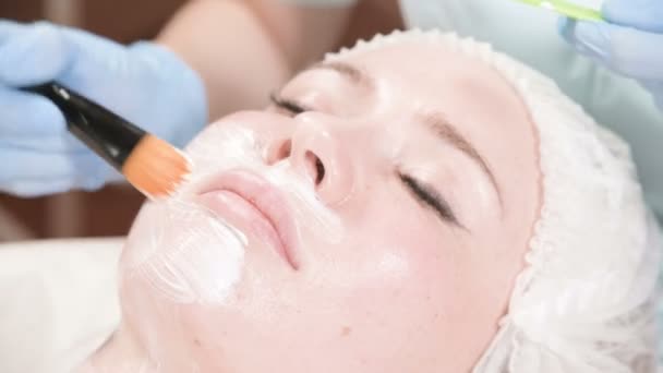 Το κορίτσι ένα cosmetologist εκτελεί τη διαδικασία καθαρισμού και ενυδάτωσης του δέρματος. Εφαρμόστε μια μάσκα με ειδικό πινέλο για το πρόσωπο μιας γυναίκας. Φροντίδα για το δέρμα σας. — Αρχείο Βίντεο
