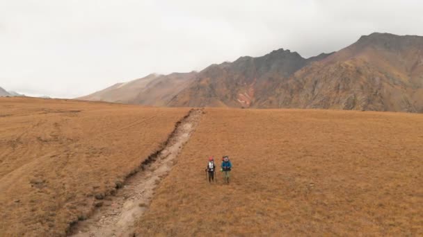 一对头戴大背包、戴着太阳镜、头戴大背包的男女旅行者沿着高山高原漫步的空中景象 — 图库视频影像