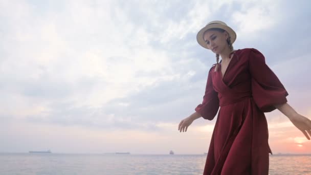 Een aantrekkelijk jong meisje in een rode zomerjurk en een strohoed kruipt voorzichtig op de stenen van de zee rotsachtige kust tegen de achtergrond van golven en zonsondergang. — Stockvideo