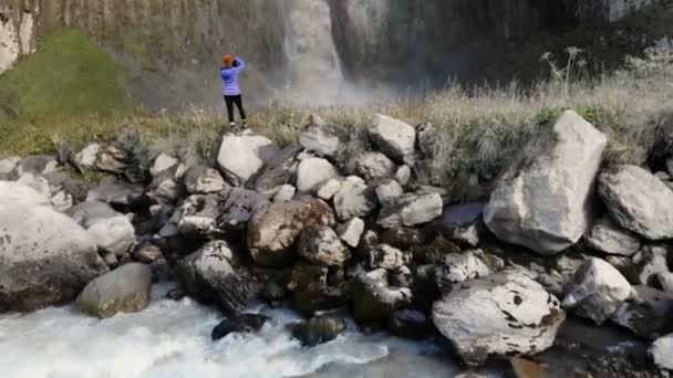 Uma menina é uma fotógrafa de paisagem profissional que está em uma pedra junto ao rio e fotografa uma grande cachoeira épica. Vista aérea. Norte do Cáucaso — Vídeo de Stock