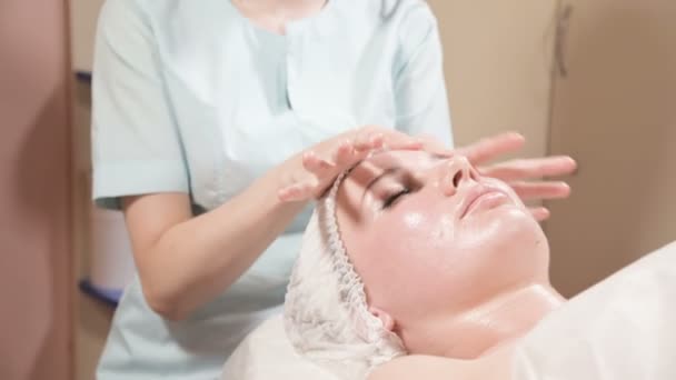 Menina esteticista em uma máscara e luvas faz uma massagem facial salubre para uma mulher atraente. Nova geração massagem cosmetologia — Vídeo de Stock