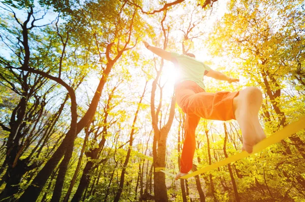 Wide angle male tightrope walker balancing barefoot on slackline in autumn forest. The concept of outdoor sports and active life of people aged