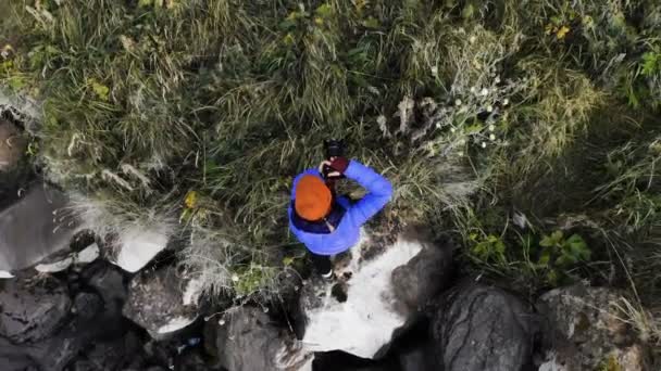 En flicka är en professionell landskapsfotograf som står på en sten vid floden och fotograferar ett stort episkt vattenfall. Flygfoto. Nordkaukasien — Stockvideo