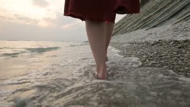 Close-up rear view of legs Caucasian girl walks along the water on the stone coast of the sea towards the setting sun at sunset with waves — Stock Video