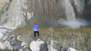 Bir kız nehir kenarındaki bir taşın üzerinde duran ve büyük destansı bir şelaleyi fotoğraflayan profesyonel bir manzara fotoğrafçısı. Hava görüntüsü. Kuzey Kafkasya