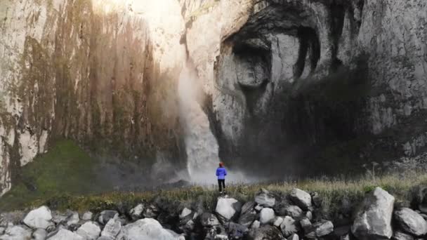 Bir kız nehir kenarındaki bir taşın üzerinde duran ve büyük destansı bir şelaleyi fotoğraflayan profesyonel bir manzara fotoğrafçısı. Hava görüntüsü. Kuzey Kafkasya — Stok video