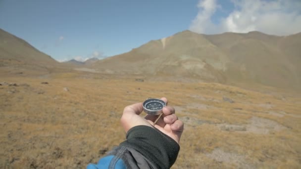 The first-person view of a male hand holding a magnetic compass rotates on the side and looking for the right direction in the background of a mountains. The concept of orienteering — ストック動画