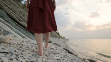 Dalgalarla kumlu kıyıya çıkan kızın yalınayak. Beyaz elbiseli Afrikalı kız gün doğumunda kıyıda deniz dalgalarında yalınayak yürüyor, yakın çekim. Yavaş çekim çekimizleme