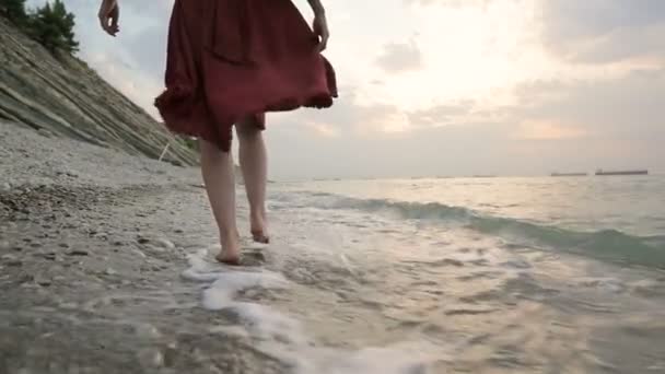 Pandangan belakang close-up gadis Kaukasia berjalan di sepanjang air di pantai batu laut menuju matahari terbenam dengan gelombang — Stok Video