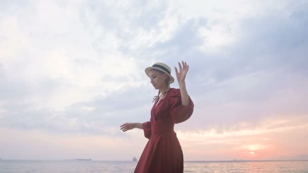 An attractive young girl in a red summer dress and a straw hat cautiously creeps on the stones of the sea rocky shore against the background of waves and sunset. — ストック動画