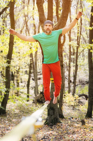 Caminhante de corda bamba macho de ângulo largo balanceando descalço no slackline na floresta de outono. O conceito de esportes ao ar livre e vida ativa de pessoas idosas — Fotografia de Stock