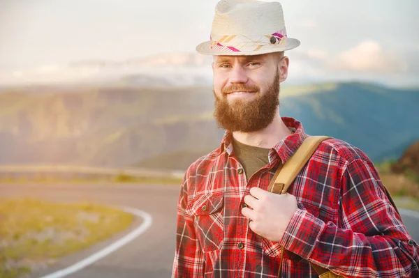 Retrato de um viajante sorridente feliz barbudo hipster com uma mochila em uma camisa xadrez e um chapéu ao lado de um carro desconhecido fica na estrada ao pôr do sol nas montanhas. Conceito de viagem feliz e confiante — Fotografia de Stock