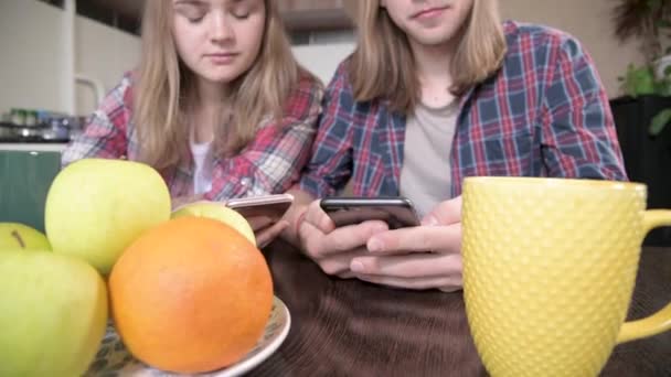 Un joven par de mellenials con el pelo largo están sentados en la mesa de la cocina con los teléfonos en sus manos. Navegación y compras en Internet desde dispositivos móviles. Concepto de familia joven moderna — Vídeos de Stock