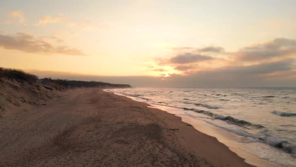 Hava aracı, gün batımında ıssız bir plajın üzerinden alçaktan uçuyor. Yuvarlanan dalgalarla kumsal kıyısı ve ufukta batan güneş. Deniz Burnu — Stok video