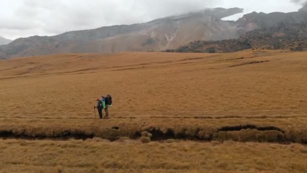 Vista aérea de un par de viajeros hombre y mujer con grandes mochilas en sombreros y gafas de sol de pie en la meseta alpina rodeado de montañas épicas — Vídeo de stock