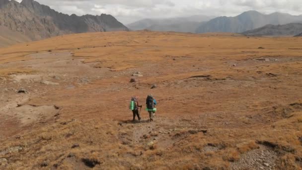 Flygfoto av ett par resenärer man och kvinna med stora ryggsäckar i hattar och solglasögon promenad längs en alpin platå omgiven av episka berg — Stockvideo