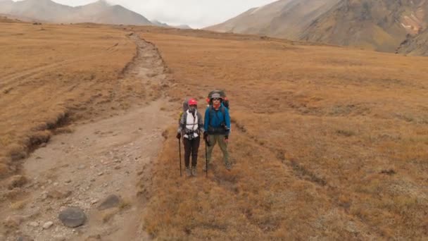 Aerial view of a couple of travelers man and woman with large backpacks in hats and sunglasses stand on alpine plateau surrounded by epic mountains — Stock Video