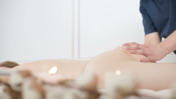 Nahaufnahme eines männlichen Physiotherapeuten, der in einem professionellen Wellness-Salon neben einem Zweig Baumwolle eine leichte Massage zum Aufwärmen der Hüften und des Gesäßes einer jungen Frau durchführt. 4k Massage Körperpflege-Konzept — Stockvideo