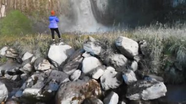 Bir kız nehir kenarındaki bir taşın üzerinde duran ve büyük destansı bir şelaleyi fotoğraflayan profesyonel bir manzara fotoğrafçısı. Hava görüntüsü. Kuzey Kafkasya