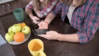 Mutfak masasında ellerinde telefonlar olan uzun saçlı bir çift tatlı kadın oturuyor. Mobil cihazlardan internet üzerinden sörf ve alışveriş. Modern genç aile kavramı