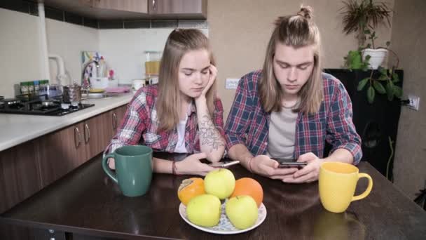 Un joven par de mellenials con el pelo largo están sentados en la mesa de la cocina con los teléfonos en sus manos. Navegación y compras en Internet desde dispositivos móviles. Concepto de familia joven moderna — Vídeos de Stock