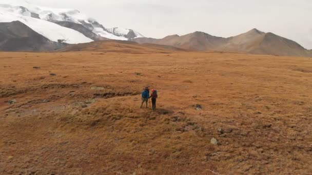 Flygfoto av ett par resenärer man och kvinna med stora ryggsäckar i hattar och solglasögon står på alpin platå omgiven av episka berg — Stockvideo