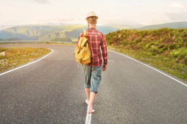 Şık yalınayak sakallı erkek otostopçu bir şapka ve bir sırt çantası ile gün batımında dağlarda bir ülke yolu boyunca yürüyor. Arkadan manzara. Paraolmadan seyahat konsepti.
