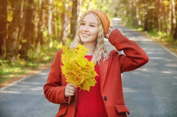 Mulher feliz atraente em polto vermelho sorrindo alegre e feliz segurando folhas de outono fora em sua mão em uma floresta de outono colorido contra o fundo da estrada . — Fotografia de Stock