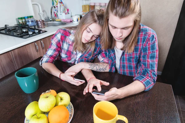 A young pair of mellenials with long hair are sitting at the kitchen table with phones in their hands. Surfing and shopping on the Internet from mobile devices. Concept of modern young family — Stock Photo, Image