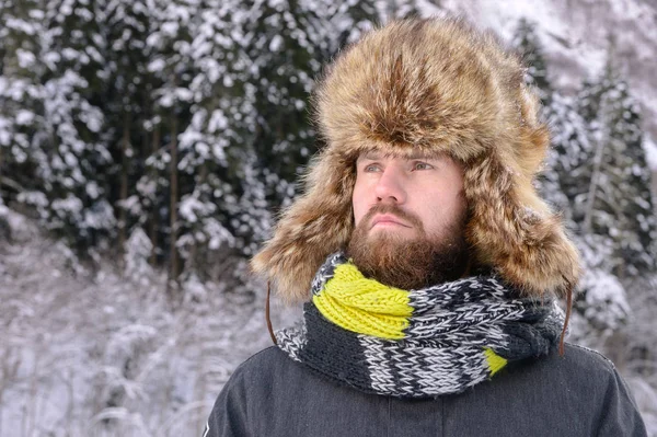 Retrato de um homem viril atraente com bigode e barba em uma floresta de inverno. Turista elegante em um chapéu de pele e um lenço brilhante — Fotografia de Stock