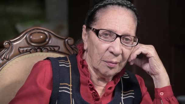 Portret van een bejaarde vrouw in een bril leunend op haar arm binnen zittend en denkend. Oudere vrouw 80 jaar oud — Stockvideo