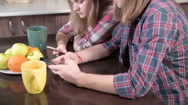 Um jovem par de mellenials com cabelos longos estão sentados na mesa da cozinha com telefones em suas mãos. Navegar e fazer compras na Internet a partir de dispositivos móveis. Conceito de família jovem moderna — Vídeo de Stock