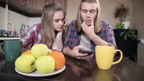 Un joven par de mellenials con el pelo largo están sentados en la mesa de la cocina con los teléfonos en sus manos. Navegación y compras en Internet desde dispositivos móviles. Concepto de familia joven moderna — Vídeos de Stock