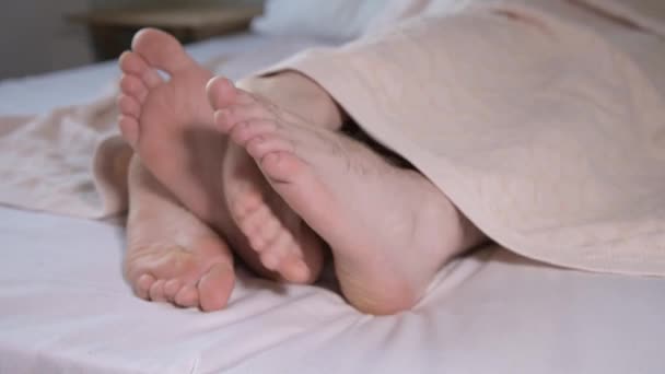 一对年轻夫妇脚的特写从卧室的被子下面伸出来。 赤脚互相爱抚，忙于打扮. — 图库视频影像