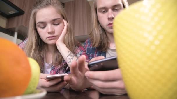 長い髪の若い2人の子午線は、彼らの手に携帯電話でキッチンテーブルに座っています。モバイルデバイスからのインターネット上でのサーフィンやショッピング。現代の若い家の概念 — ストック動画