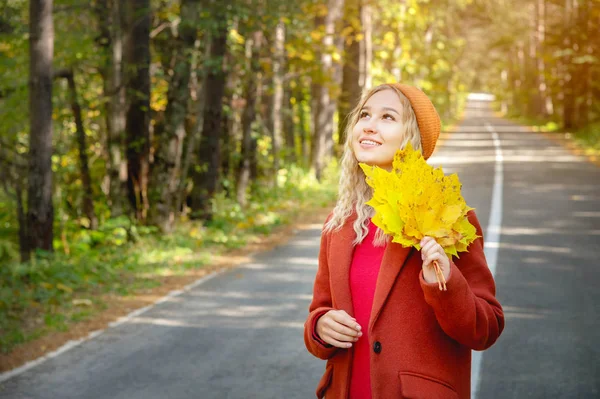 Mulher feliz atraente em polto vermelho sorrindo alegre e feliz segurando folhas de outono fora em sua mão em uma floresta de outono colorido contra o fundo da estrada . — Fotografia de Stock