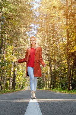 Kırmızı ceketli, neşeli ve mutlu bir kadın elinde sonbahar yapraklarıyla asfalt yolda yürüyen renkli bir sonbahar ormanında elini tutuyor..