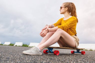 Güneş gözlüğü ve şort kolunda bir dövme ile Çekici genç kız gri gökyüzü bir arka plan üzerinde bir banliyö park onu longboard oturur. Millennials nesil ve eğlence kavramı