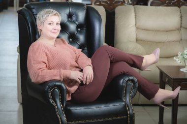 Orta yaşlı bir kadının portresi. Mobilya işinin sahibi ve yöneticisi pahalı bir deri koltukta oturuyor ve gülümsüyor. Kestirme yol güçlü kadın.