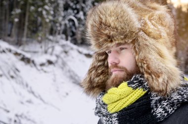 Kış ormanında bıyıklı ve sakallı çekici bir adamın portresi. Kürk şapkalı şık bir turist ve parlak bir eşarp.