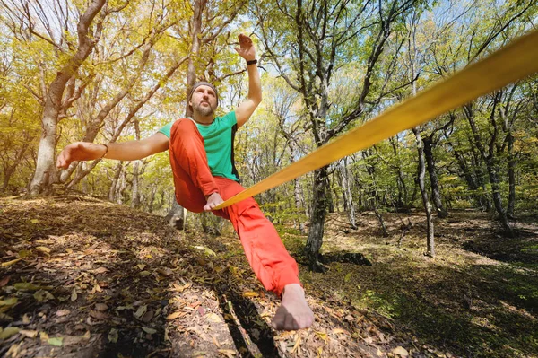 Wide angle male tightrope walker balancing while sitting barefoot on slackline in autumn forest. The concept of outdoor sports and active life of people aged
