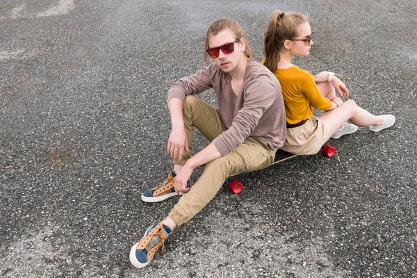Um jovem casal atraente de óculos de sol se senta de costas para trás em seu longboard em um estacionamento suburbano. O conceito de uma geração jovem da família millennials estilo e lazer — Fotografia de Stock