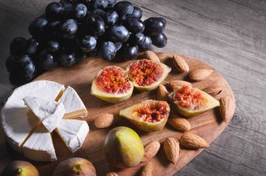 Ahşap bir kesme tahtası üzerinde Natürmort peynir plaka kabuk kalıp badem ve siyah üzüm bir demet yanında dilimlenmiş incir ile dilimlenmiş peynir yatıyor. Lezzetli ve sağlıklı gıda konsepti