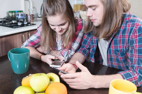 A young pair of mellenials with long hair are sitting at the kitchen table with phones in their hands. Surfing and shopping on the Internet from mobile devices. Concept of modern young family — Stock Photo, Image
