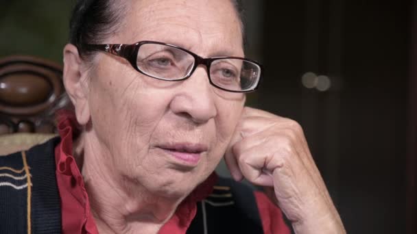 Portret van een bejaarde vrouw in een bril leunend op haar arm binnen zittend en denkend. Oudere vrouw 80 jaar oud — Stockvideo